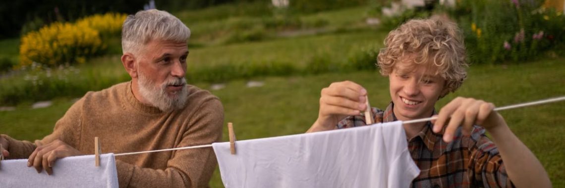 Elderly man and teenage boy joyfully hanging laundry in a sunny garden setting.